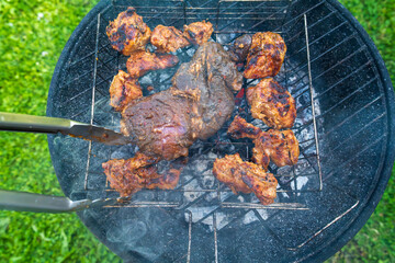 Grilled pork pieces and steak are cooking on a barbecue grill over hot coals. A person using metal tongs to flip meat during an outdoor summer cooking session.