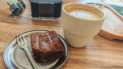 Cup of cappuccino with a piece of chocolate brownie on metal plate at wooden table. This image...