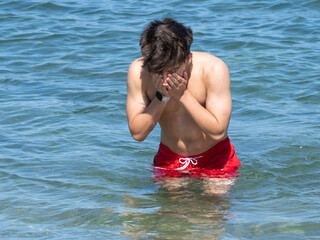 Young man is refreshing himself by washing his face in the ocean on a sunny summer day