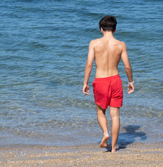 Teenager walking into the sea water wearing red swimming trunks on a summer day at the beach
