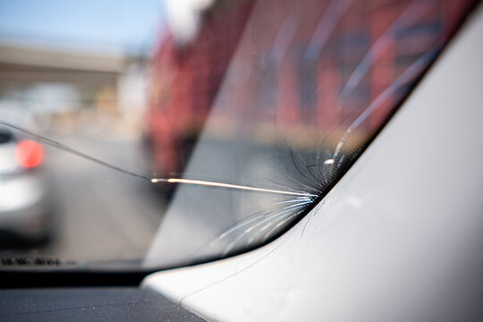 A close-up view of a cracked windshield caused by a small rock while driving on a crowded highway under bright daylight.