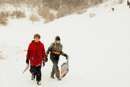 Kids hike up snowy hill carrying sleds during snowfall