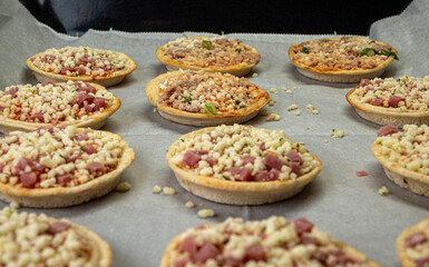 A tray of mini pizzas with cheese and meat toppings. The pizzas are arranged in a neat row on a baking sheet