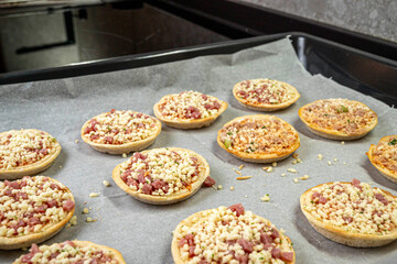 A tray of mini pizzas with cheese and meat toppings. The pizzas are on a baking sheet and are ready to be baked