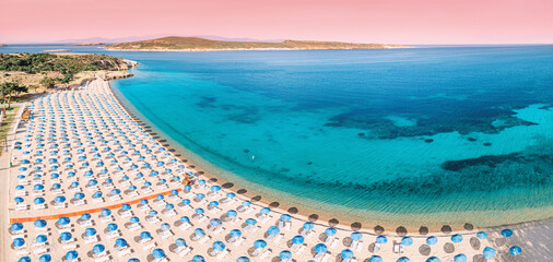 Rows of beach umbrellas line a pristine sandy shore, meeting the crystal clear turquoise waters of a picturesque bay under a soft pink sky
