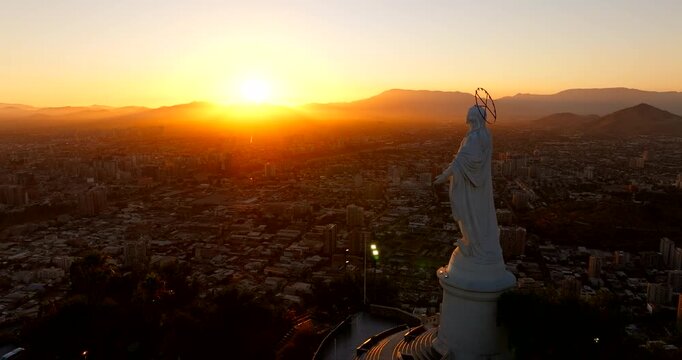 Virgin Mary statue on Cerro San Cristobal hill, overlooking Santiago cityscape at golden hou, sun behind Andes mountains, Chile. Aerial forward