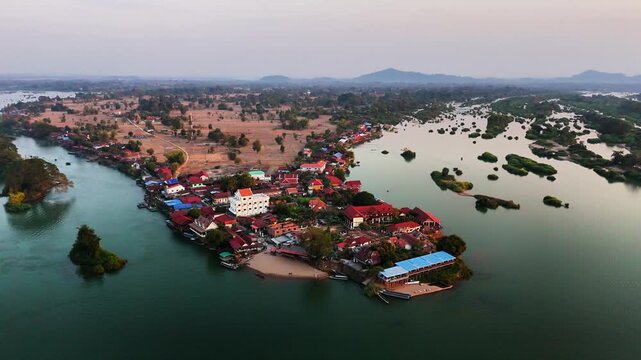 Aerial drone orbit reveal core of Don Det village in Laos, riverside stilted bungalows, historic wooden bridge linking to Don Khon, and the small Buddhist temple along the Mekong shoreline at sunset