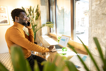Smiling African American man wearing glasses and sweater working on laptop while talking on smartphone