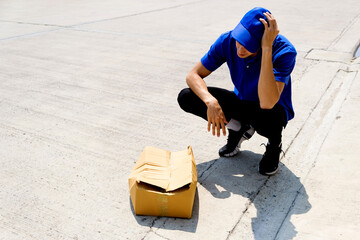 Courier in blue uniform squats beside a crushed cardboard box on the street, holding his head in...