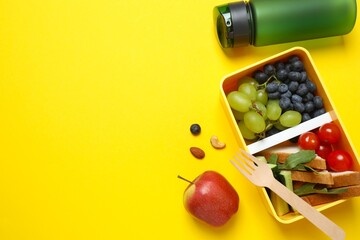 Top view photo of lunch box with healthy snacks, fruits and  bottle of water on a yellow background