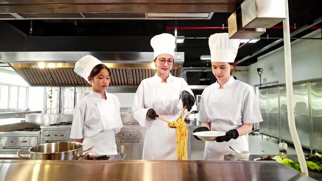 Chef instructor guides two young culinary students in plating pasta during a professional cooking class in a commercial kitchen, emphasizing hands-on learning, food preparation, and teamwork skills.