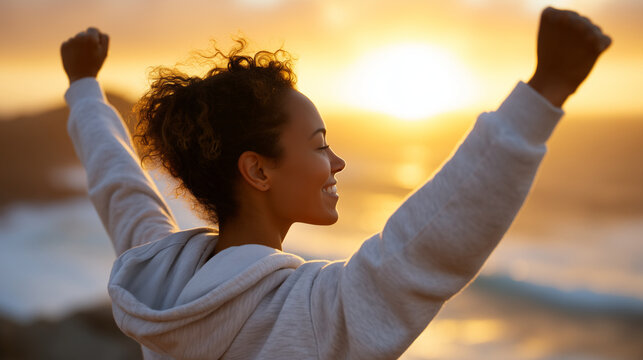 Woman in sportswear with raised fists facing the ocean sunrise, backlit by golden light, symbol of strength, resilience, morning exercise and personal growth morning sunlight, silh - Powered by Adobe