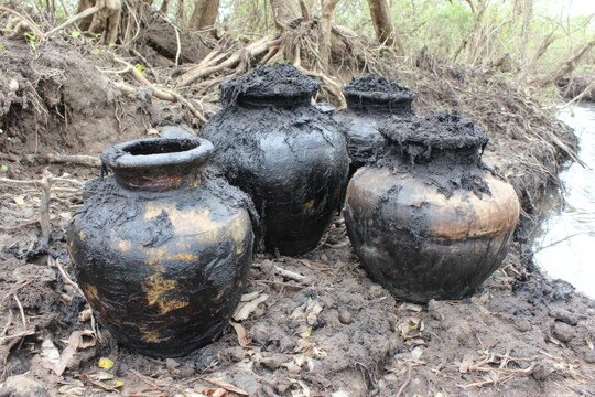 Several large, dark-coated earthenware jars sit on muddy ground near a waterway