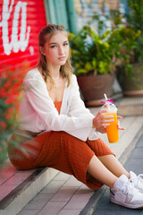 A beautiful teenage model girl posing on the streets of old town during street style photo shooting drinking ice fruit beverage