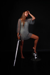 Beautiful warrior woman in chainmail holding sword and standing by her bare foot on helmet full...