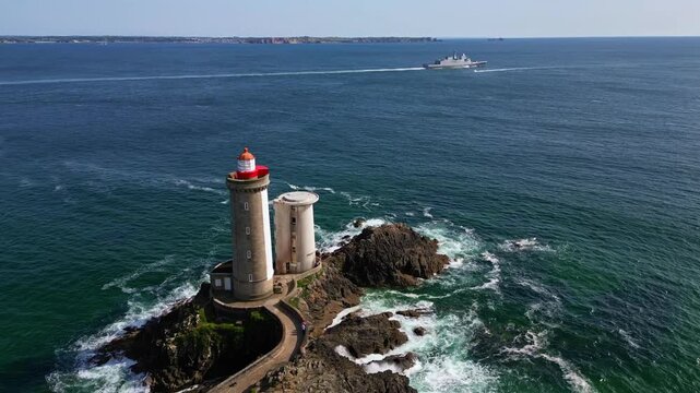 Drone orbiting around Petit Minou lighthouse in Brittany, revealing the rocky shore and path as a military ship crosses the open ocean in the background - Plouzan&eacute;, France