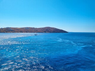 A bright blue sea with a small sailing boat off the coast of a Rocky island under a clear summer...