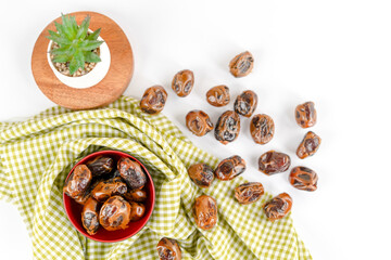 A red bowl filled with dried dates placed on a green checkered fabric, surrounded by scattered...