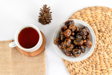 A white bowl filled with dried dates paired with a cup of tea, placed on woven and burlap mats, with a pine cone on a white background.