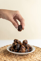 A hand picking up a date from a small plate full of dates, presented on a woven placemat with soft daylight and a warm beige background.