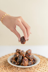 A hand picking up a date from a small plate full of dates, presented on a woven placemat with soft daylight and a warm beige background.