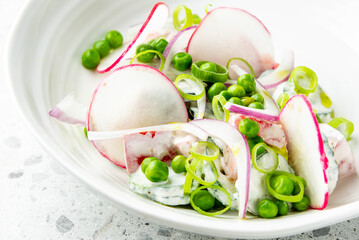 Fresh salad with sliced radishes, green peas, and spring onions in a white bowl on a light surface,...