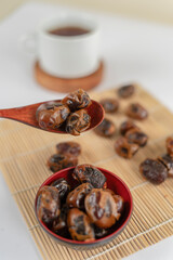Three dried dates resting on a wooden spoon, with a red bowl full of dates below and a cup of tea in the background on a bamboo mat.