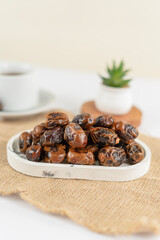 A neat pile of dried dates placed on a marble tray atop a burlap mat, with a cup and a small plant blurred in the background, under soft natural lighting.