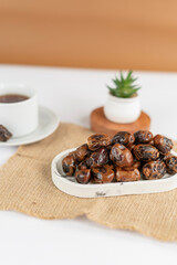 A neat pile of dried dates placed on a marble tray atop a burlap mat, with a cup and a small plant blurred in the background, under soft natural lighting.