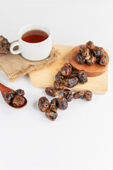 A scattered arrangement of dried dates on a white surface, accompanied by a wooden spoon and a cup of tea, all set on a rustic wooden board and burlap mat