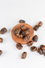 A round wooden plate filled with dried dates, surrounded by scattered dates on a clean white surface, arranged in a simple and minimal layout.