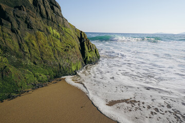 Waves washing onto a sandy beach with moss-covered rocks at the shoreline.