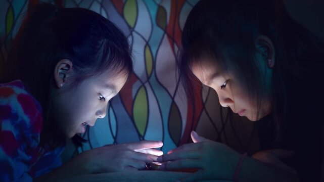 Two Asian child lying in bed using smartphone at night, screen light illuminating face, concept of screen time and digital addiction in kids