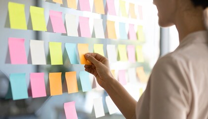 A businesswoman organizes tasks by placing a colorful sticky note on a glass wall during a brainstorming session.