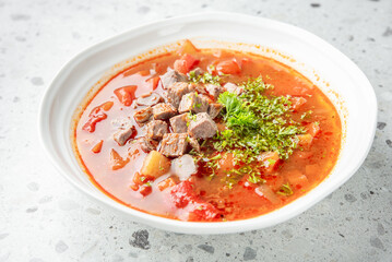 Homemade beef soup with tender meat, potatoes, and vegetables in tomato broth, garnished with parsley, served in a white bowl on a speckled countertop.