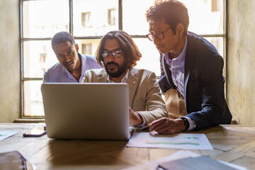 Diverse business team working together on laptop in a modern office meeting. Multi-ethnic male colleagues brainstorming a strategy. Teamwork, collaboration, startup culture, and inclusion concepts.