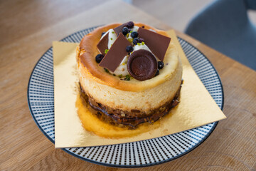 Close-up of delicious baked cheesecake with cream topping, chocolate decorations and blueberries on a gold base. Served in cafe or home setting, perfect for menu, bakery, or dessert promotion.
