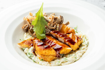 Grilled chicken breast with teriyaki sauce served on creamy risotto, garnished with fried mushrooms and herbs, plated on white porcelain in a gourmet restaurant setting.