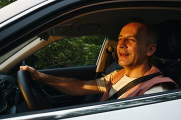 Naklejka premium Middle aged Caucasian man driving car during family road trip, smiling and looking ahead with one hand on steering wheel, sunlight illuminating face, seatbelt fastened