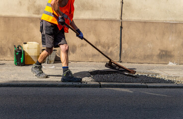 Road worker in a reflective vest leveling asphalt during street surface repair. Manual labor on a...