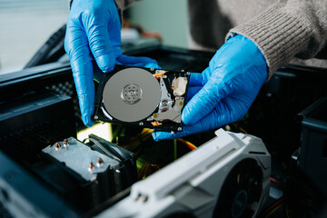 Closeup of gloved technician holding exposed hard drive above desktop PC. Ideal for data recovery,...