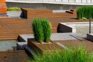 Modern public space with wooden terraces, ornamental grasses in planters, and elements of small...
