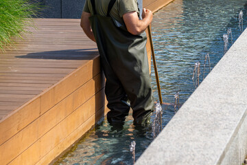 Worker in waterproof overalls cleaning a modern city fountain. Wooden terraces and ornamental...