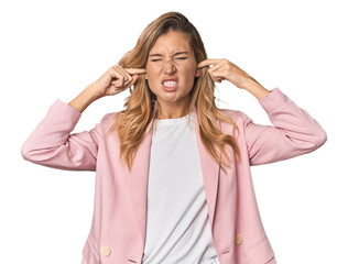 Young blonde in stylish pink suit covering ears with fingers, stressed and desperate by a loudly...