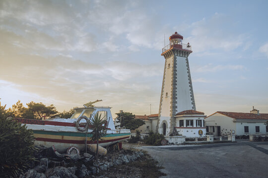 View of an old boat with peeling paint sits beside a tall white lighthouse under a soft sky, as the sun sets, casting a warm glow, Leucate, Occitanie, France.
