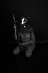 Obraz premium Female knight in chainmail and helmet holding sword and sitting on her knees low key black and white portrait