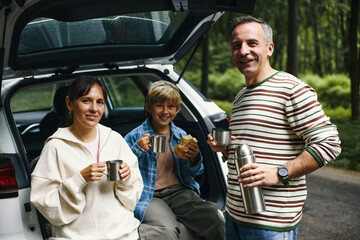 Caucasian middle aged man, Caucasian woman, and Caucasian boy sitting at open car trunk drinking...