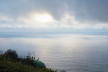 Panoramic view of a serene ocean with the sun breaking through a cloudy sky, seen from a grassy cliff edge, creating a calming atmospheric landscape.