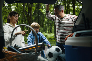 Caucasian middle aged woman, Caucasian middle aged man, and Caucasian boy unloading car trunk together during family road trip, visible picnic basket, cooler, and soccer ball in foreground