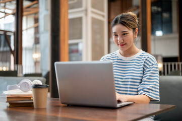 Asian young adult woman looking and using laptop aside books and coffee sits at wooden table in cafe